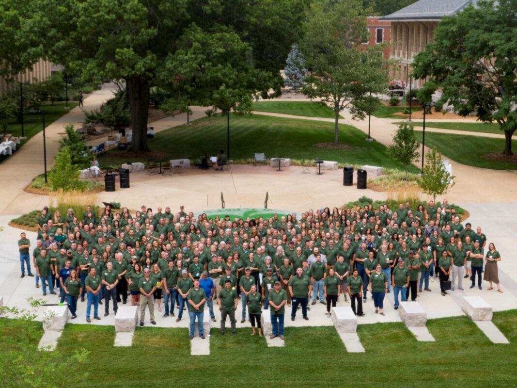 A large group of people wearing matching green shirts, posed together outdoors on a paved area surrounded by grass, trees, and a campus-like setting.